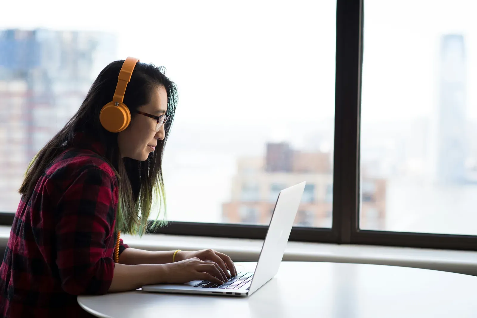 A student in headphones working on a laptop by a window overlooking a city